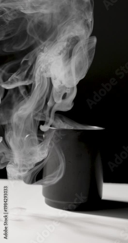 A matte black mug emits swirling white steam against a dark background, lit with dramatic contrast on a minimal surface, creating a moody, atmospheric scene with copy space