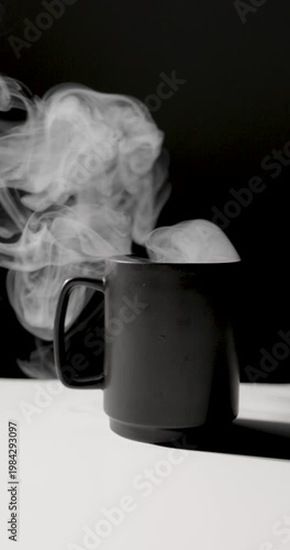 A matte black mug emits swirling white steam against a dark background, lit with dramatic contrast on a minimal surface, creating a moody, atmospheric scene with copy space