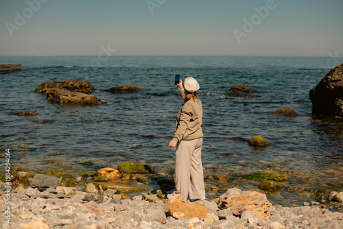 woman from behind taking photo of sea standing on rocky beach in sunny day