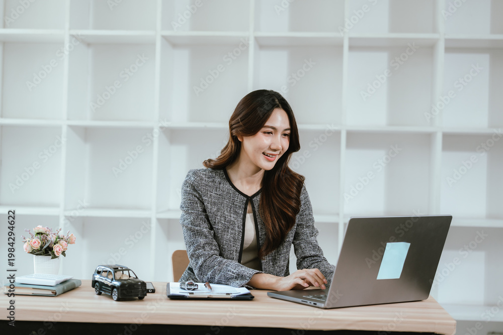 Fototapeta premium A smiling businesswoman working on a laptop at a desk with a car model and documents, representing auto business, insurance, and modern office productivity.