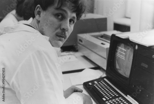 Young man in white lab coat sitting at early computer terminal with keyboard and CRT monitor in laboratory, scanned black and white print circa 1980s
