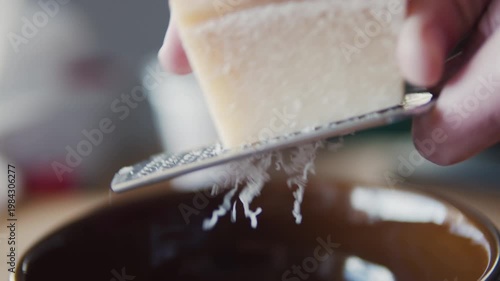 Close-up cheese being grated on metal grater into brown plate. Food preparation, cooking process, macro kitchen shot, fresh shredded cheese texture.