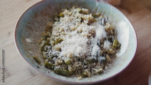 Hand sprinkling grated cheese over risotto in bowl on wooden table. Italian food preparation, cooking process, creamy dish finishing, close-up.