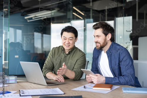 Two diverse male colleagues are smiling while discussing a project, looking at a laptop, and collaborating with a tablet in a modern office environment