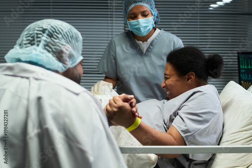 Obraz na plátně Woman gripping partner hand while laboring in hospital bed, nurse assisting childbirth