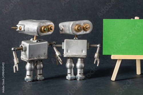 Metal silver robots at wooden easel and empty green placard. Notice board concept, promotional flyer, event announcement leaflet