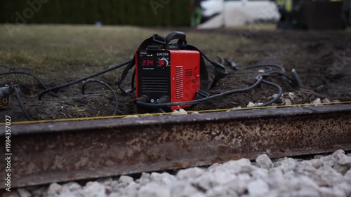 Red EW256 welding inverter shows 234 beside rusty steel rail and gravel. Cables move slightly as placement adjusts. Low angle, shallow depth, late day light.