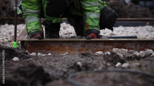 Gloved worker kneels, adjusts taut yellow line on rusted rail, grips hammer, with tape on rebar and bottle marker visible, shallow focus, overcast light.