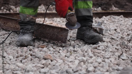 Railway maintenance worker grinds rusted rail, sparks fly at daytime. Lone technician cuts and smooths metal, cable trails across ballast, gritty shallow depth of field.