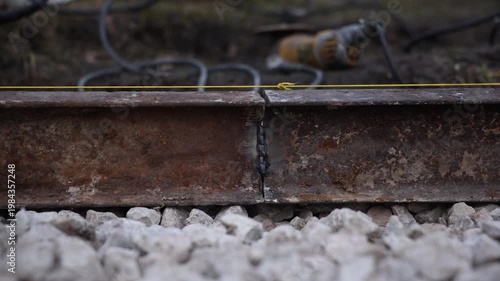 Rusted steel rails join with uneven weld beads as yellow twine marks a guide. Coarse ballast lies ahead, a power drill and cords sit blurred, natural light reveals wear.