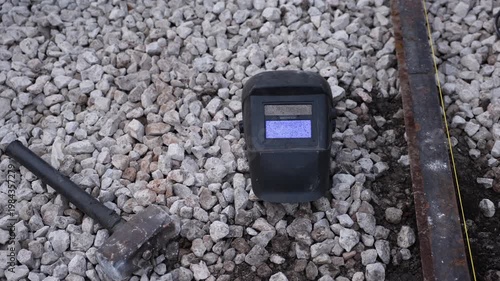 Welding helmet on crushed stone beside sledgehammer and steel rail. Taut yellow string line indicates railwork. Muted metal tones and gravel suggest recent alignment.