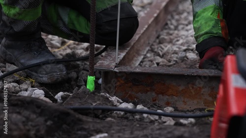 Two workers kneel on gravel ballast and weld a rusted rail as sparks fly, neon green tape marks rebar, red gloves and scuffed boots show on site repair work