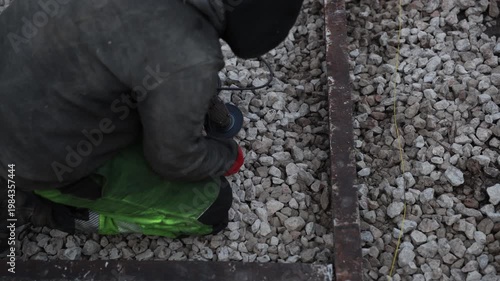 Kneeling worker uses angle grinder on rusty beam set in crushed stone. Yellow guideline runs parallel. Red gloves and green knee pads stand out in muted winter light.