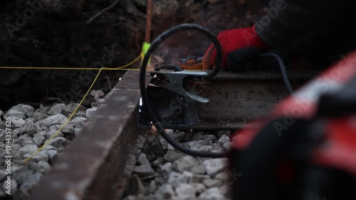 A gloved hand operates a grinder on a rusted steel rail as sparks fly. A clamp and heavy cable rest on crushed ballast. Yellow guide line and weld bead are visible.