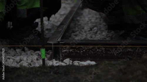 Two workers kneel on crushed stone ballast, align a rusted I beam rail with a taut yellow guide string, sparks flicker as a thin rod suggests welding or grinding.