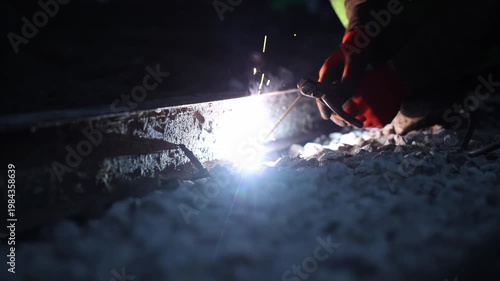 Title  Night rail welding with red gloves and sparks in shallow focus. Technician holds torch to railroad rail, arc throws sparks and smoke over gravel, cool grading, slow shutter.