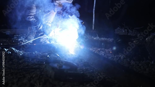 Welder kneels on rail as arc weld sparks and smoke rise at night. Title appears here. Hand moves, molten metal flows, gravel ballast and tools sit near, high contrast cool cast.