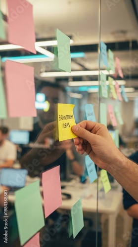 A hand places a yellow sticky note saying brainstorm ideas on a glass wall. Professional office setting focused on creativity. Ideal for business, teamwork, and planning concepts.