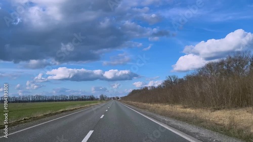 View from a car window of a straight road with white lane markings under a blue sky with clouds. Concept of travel, journey, movement forward, and freedom. Transportation, road trip, and perspective.