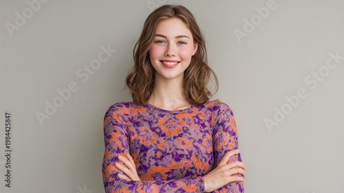 Smiling young woman with brown shoulder-length hair wearing colorful floral mesh top, arms crossed against grey wall