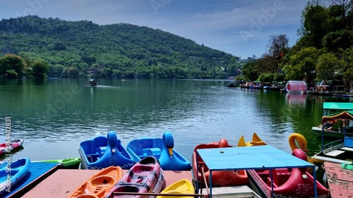 A serene lakeside view with colorful kayaks and swan-shaped paddle boats resting at dock, overlooking a wide calm lake with a lone boat drifting toward a lush green hillside under a clear sky.