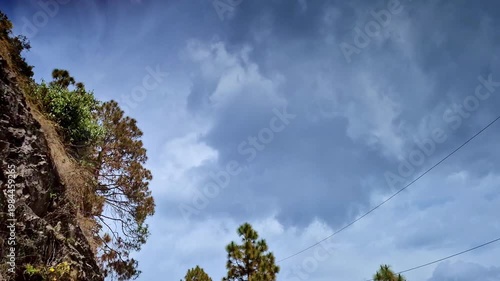 A steep rocky cliff in Mukteshwar, Uttarakhand rises sharply with patches of grass and shrubs clinging to its surface, set against a wide sky filled with thick, drifting clouds.