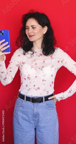 Happy young woman with curly hair taking selfie and recording video on blue smartphone over red background. Beautiful girl in floral sheer blouse smiling and posing for mobile phone camera indoor