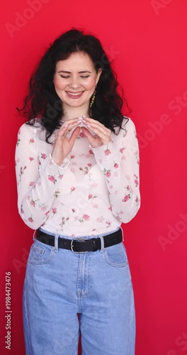 Cunning and playful young woman winking and plotting something with hands gesture on red background. Cheerful brunette girl in floral blouse looking at camera with sneaky and joyful expression