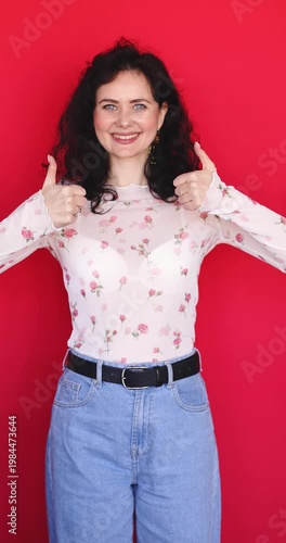 Happy young woman showing thumbs up with both hands and laughing on red background. Joyful brunette girl in floral sheer blouse giving double like gesture with positive facial expression in studio