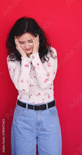 Stressed and upset young woman covering face with hands and looking disappointed on red background. Frustrated brunette girl in floral blouse expressing regret, shame or failure in studio