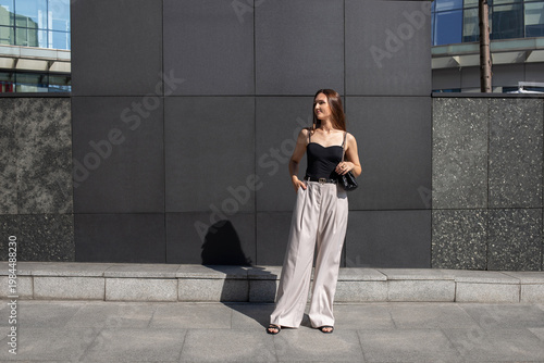 Young beautiful brunette woman wearing beige palazzo pants, black tank top, heeled sandals and small bag standing outdoor on a city street against black wall. Trendy casual outfit. Street fashion.