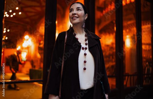 Foto Confident mature Latin woman walking in night city with blurred golden lights ba