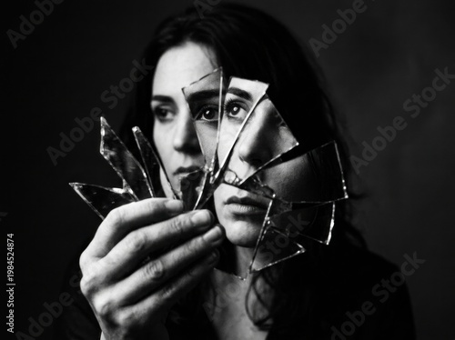 Black and white portrait of woman holding shattered mirror fragments reflecting fragmented face.
