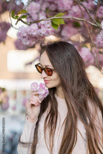 Woman with long brown hair wearing sunglasses poses among blooming sakura cherry blossom trees in a vibrant spring garden, soft focus background enhancing floral colors