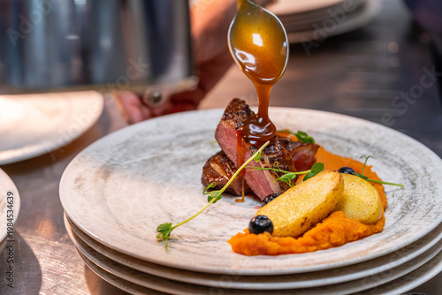 Chef pouring sauce over gourmet meat dish with vegetables in professional kitchen