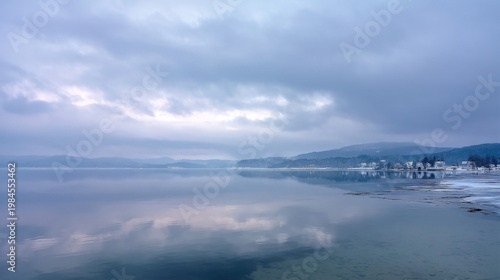 Overcast sky and mirrored lake reflect distant mountains with a snowy lakeside town shrouded in mist.