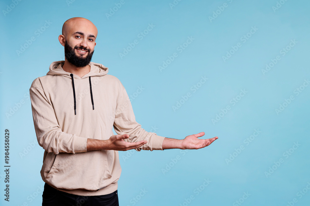 Naklejka premium Cheerful smiling arab man pointing to left while looking at camera. Happy joyful young person advertising product and showing to side portrait while posing on blue background