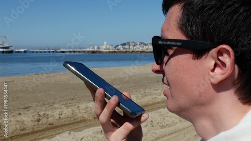 Young man recording voice message on smartphone at the beach.