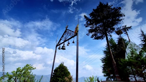 A serene hilltop scene in Mukteshwar, Uttarakhand features a decorative metal frame with hanging temple bells silhouetted against a vivid blue sky, surrounded by tall pine trees and distant mountains.