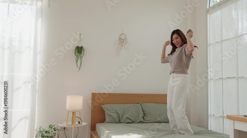 Wide shot of happy Asian woman dancing on bed listening to music