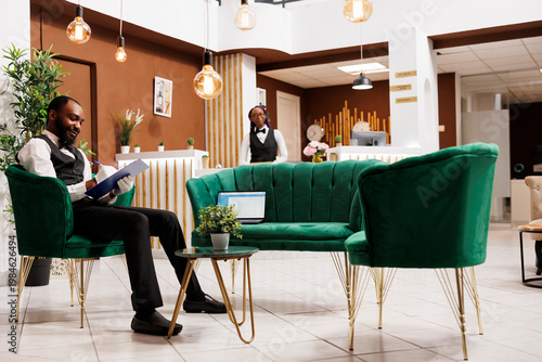Young African American smiling man bellboy wearing uniform and white gloves sitting at hotel lounge area holding clipboard making notes. Bellhop filling information of luggage in arrival errand card