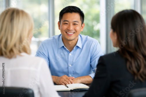 Smiling Asian businessman in a light blue shirt, confidently engaging in a professional meeting. Happy male executive, actively listening and collaborating with two female colleagues. Diverse