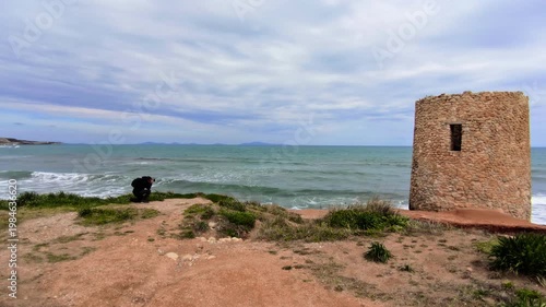 Rear view of a kneeling photographer capturing a historic stone watchtower during a sea storm, with crashing waves and a dark grey sky over the coast.