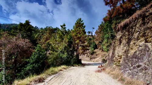 A winding dirt road in Mukteshwar, Uttarakhand passes through dense pine forest, with rocky cliffs on one side and lush greenery under a dramatic cloud-filled sky.