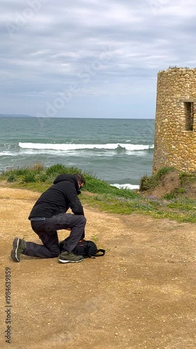 Cinematic slow motion rear view of a kneeling photographer on a rugged coastal path as he opens his backpack and carefully removes his reflex camera with the scenic Mediterranean sea in the background