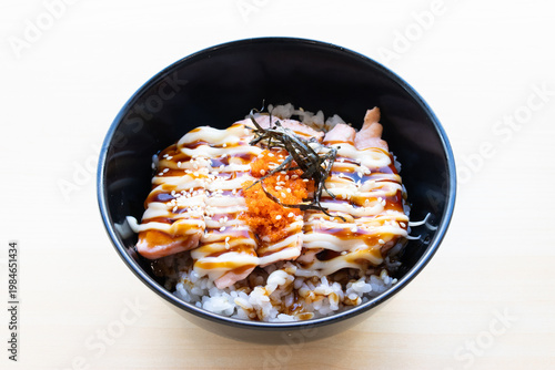 Top view of salmon donburi, grilled salmon with mayonnaise, sweet sauce, and ebiko on rice in a black bowl on a wooden table.