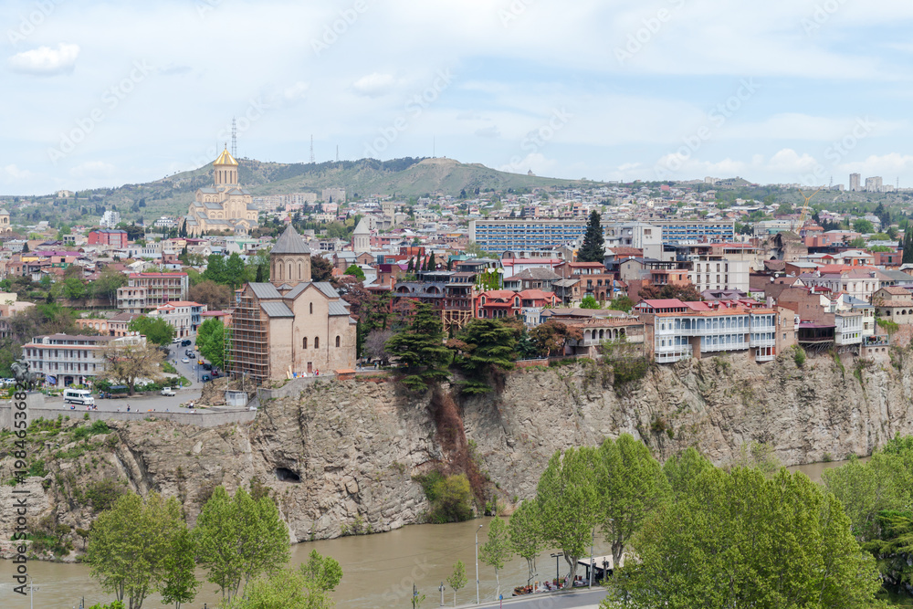 Obraz premium Panoramic view of Tbilisi featuring the historic Metekhi Church on a rocky cliff and the golden-domed Sameba Cathedral in the background, with riverfront trees, colorful houses and urban hillside