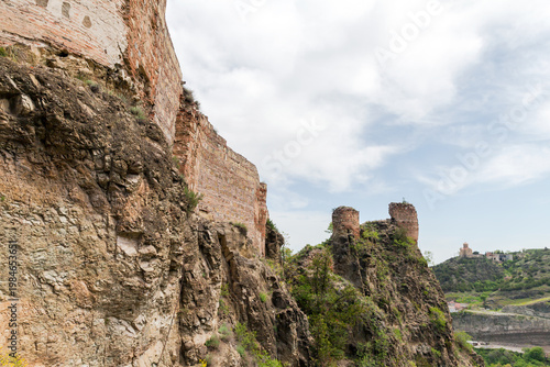 Tbilisi, Georgia. Ancient Narikala fortress ruins cling to a steep rocky cliff above a green valley, evoking history, adventure, and dramatic natural beauty beneath a soft, cloudy sky