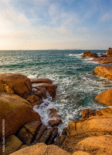 Scenic view of the Atlantic Ocean waves crashing against pink granite rocks on the famous Pink Granite Coast in Brittany, France. Warm evening light enhances the natural colors of the rugged shoreline