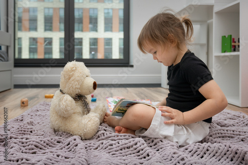 little girl reading a book on the floor in her children's room with a toy teddy bear
